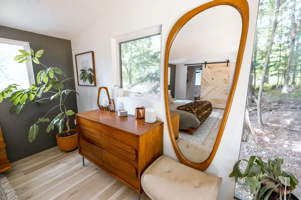 A bedroom with a wooden mid-century dresser, a tall wood-framed mirror and houseplants