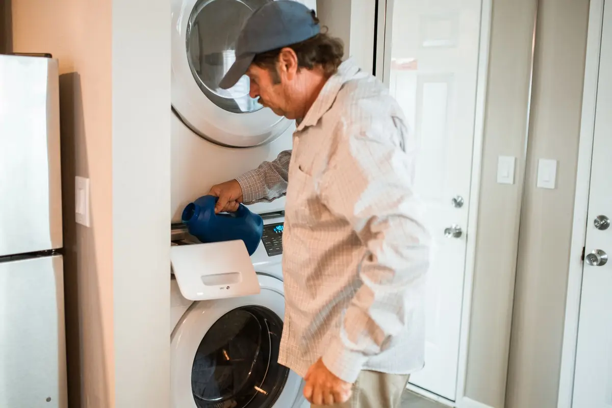 A man adding detergent to a stacked washer and dryer in a small laundry room