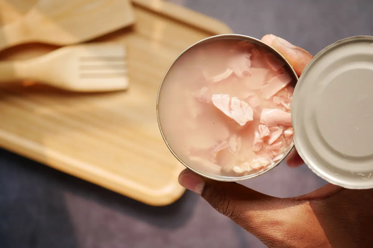 Hands holding an opened can of tuna and its lid over a wooden tray
