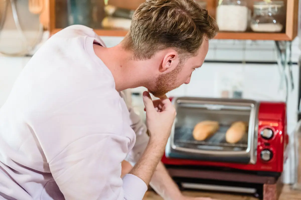 A man watching bread rolls bake in a small countertop oven