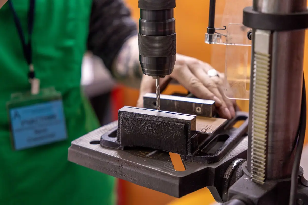 A hand lowering a drill press bit into a workpiece clamped in a vise