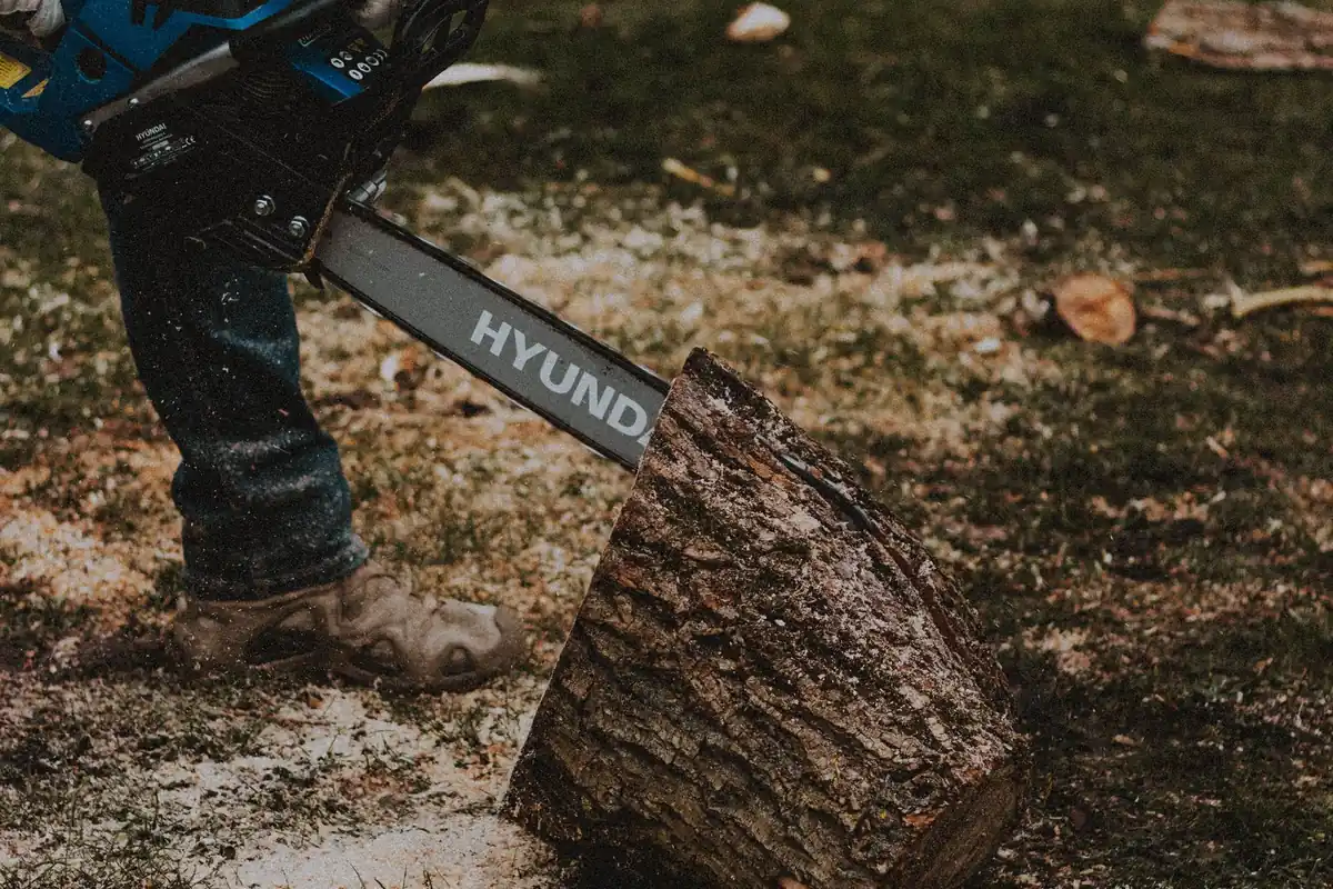 A gloved worker cutting a log with an electric chainsaw next to a firewood pile
