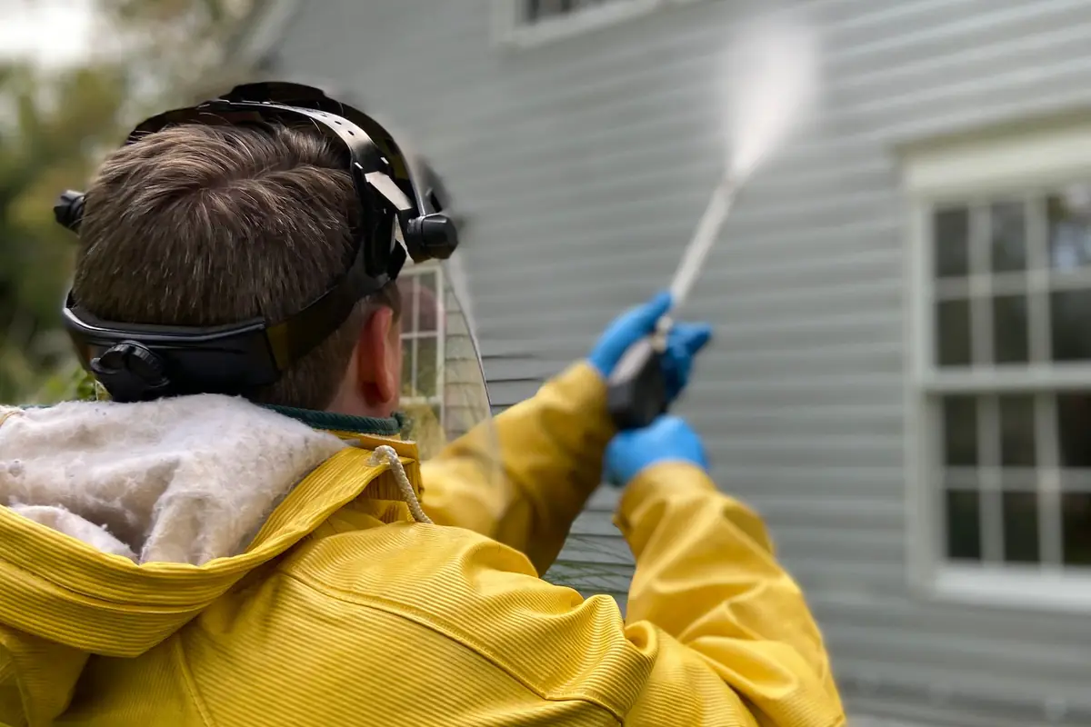 A person in a yellow raincoat and ear protection pressure washing house siding