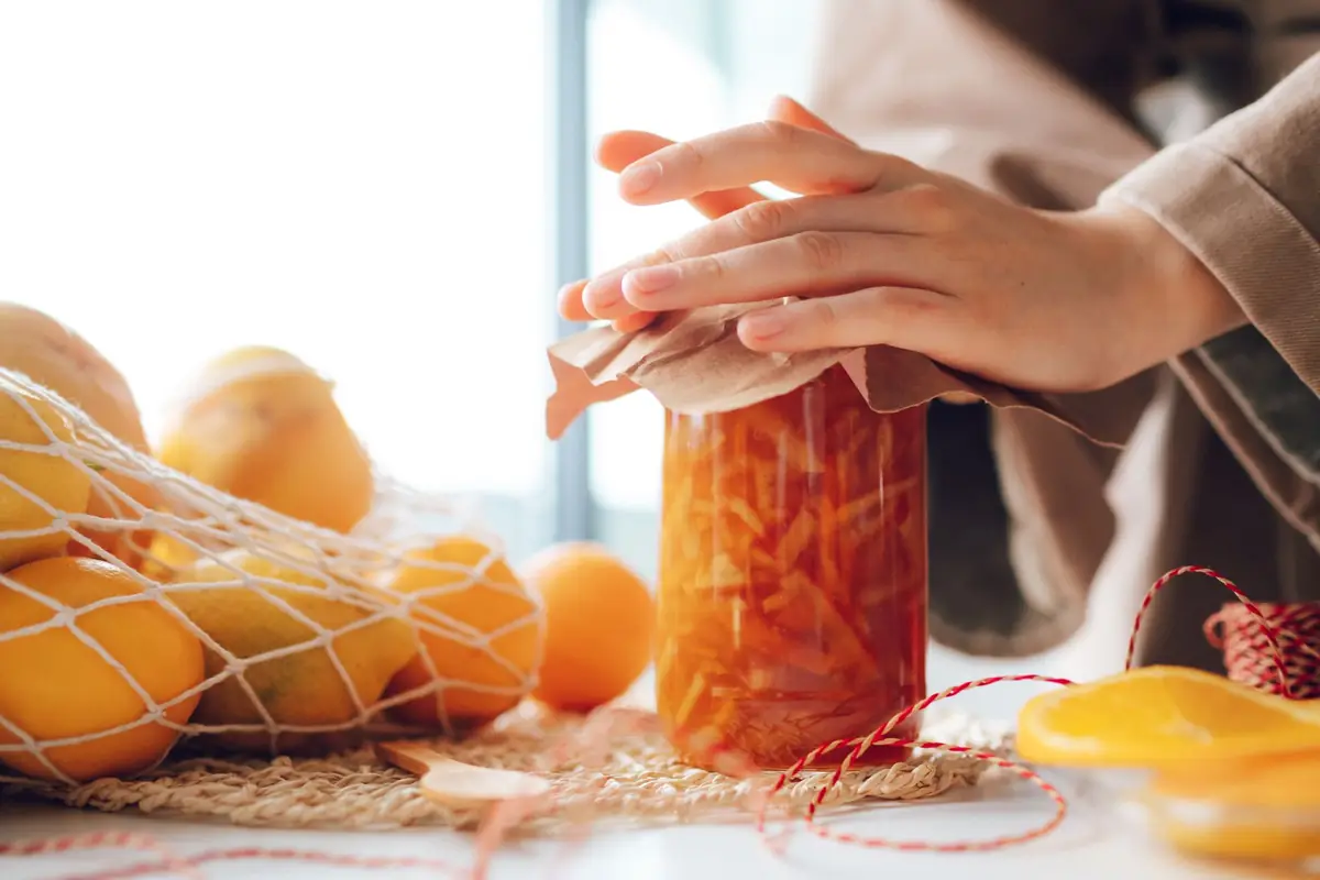A hand twisting the lid of a jar of orange marmalade beside fresh oranges