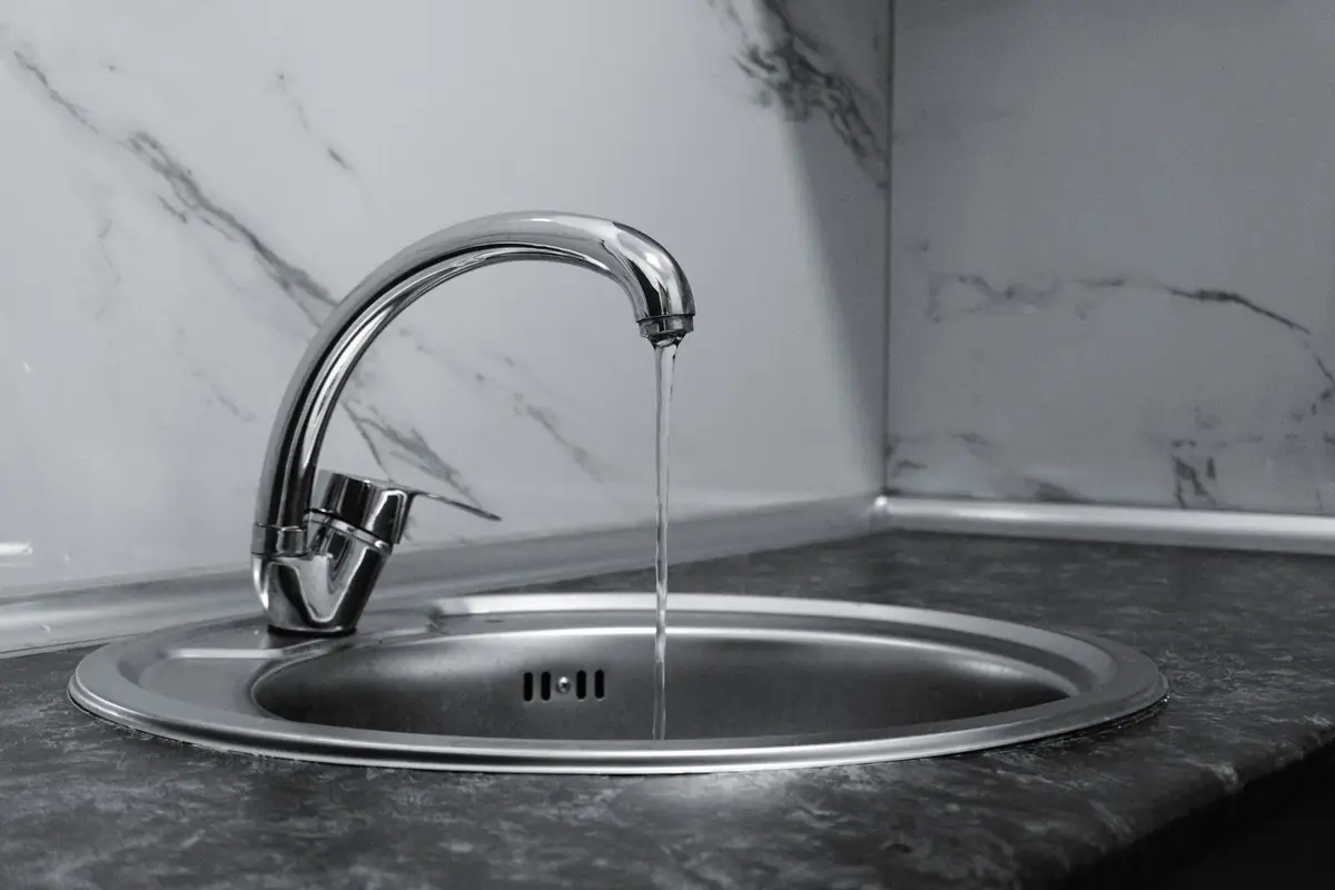 A chrome faucet running into a round stainless sink against a marble wall