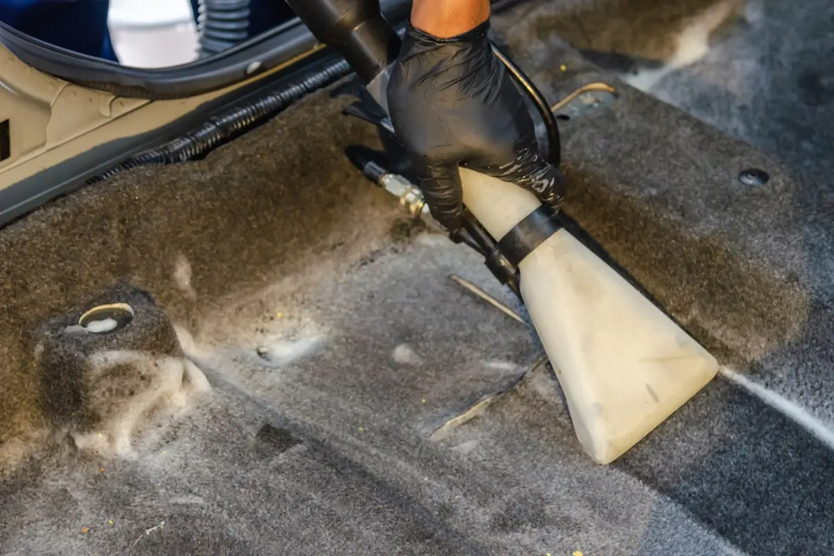A gloved hand vacuuming the carpet of a car with a crevice nozzle