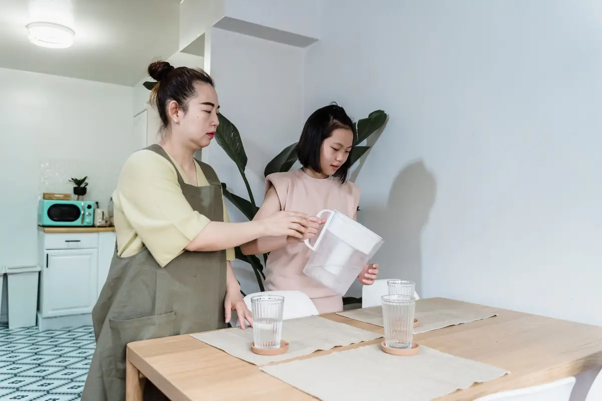 A woman pouring water from a pitcher into glasses at a kitchen table