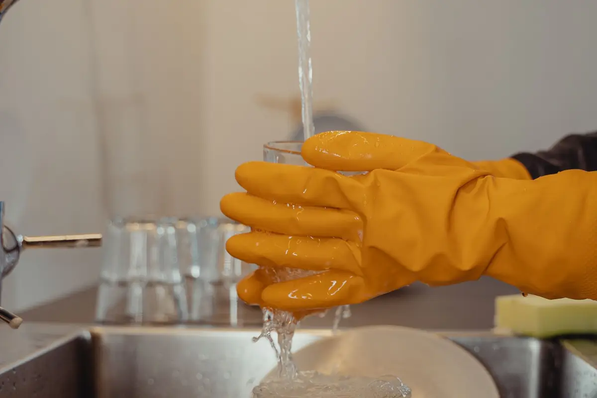 Gloved hands rinsing a glass under running water at a kitchen sink
