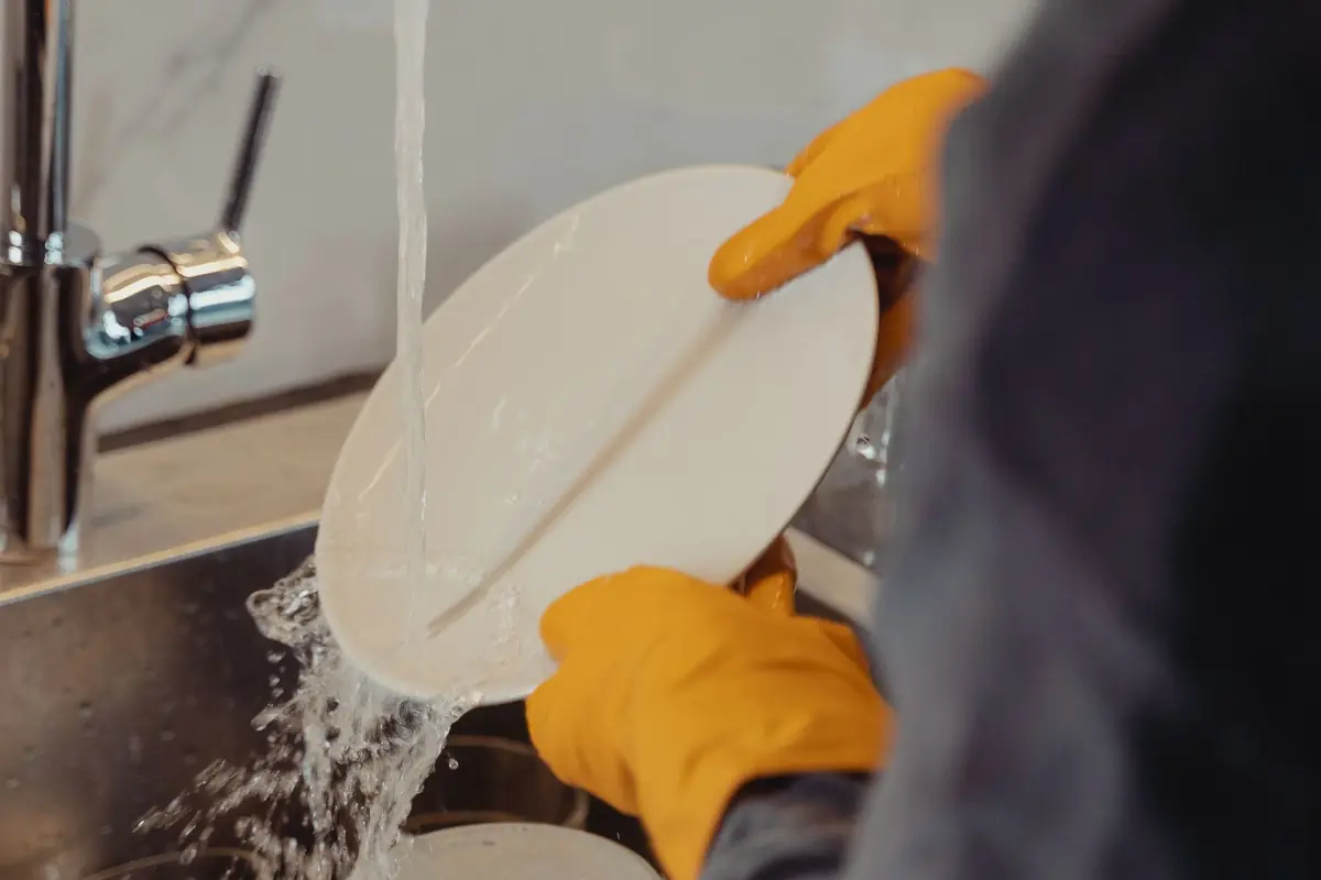 Gloved hands rinsing dishes under a kitchen faucet