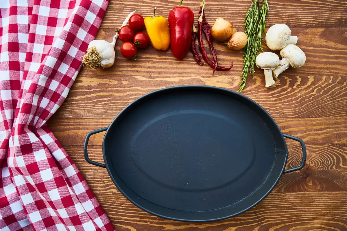 An empty black cast iron pan on a wooden table with a red checked cloth and fresh vegetables