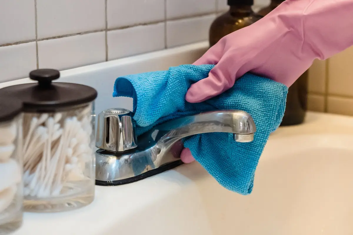 A gloved hand wiping a chrome faucet with a blue microfiber cloth