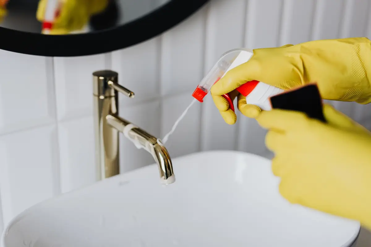 Yellow-gloved hands spraying cleaner on a brass faucet