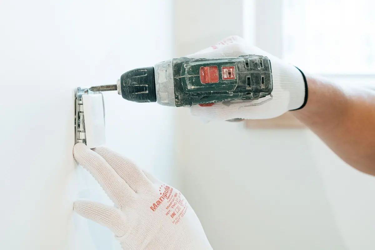 A gloved hand holding a cordless drill against a white wall